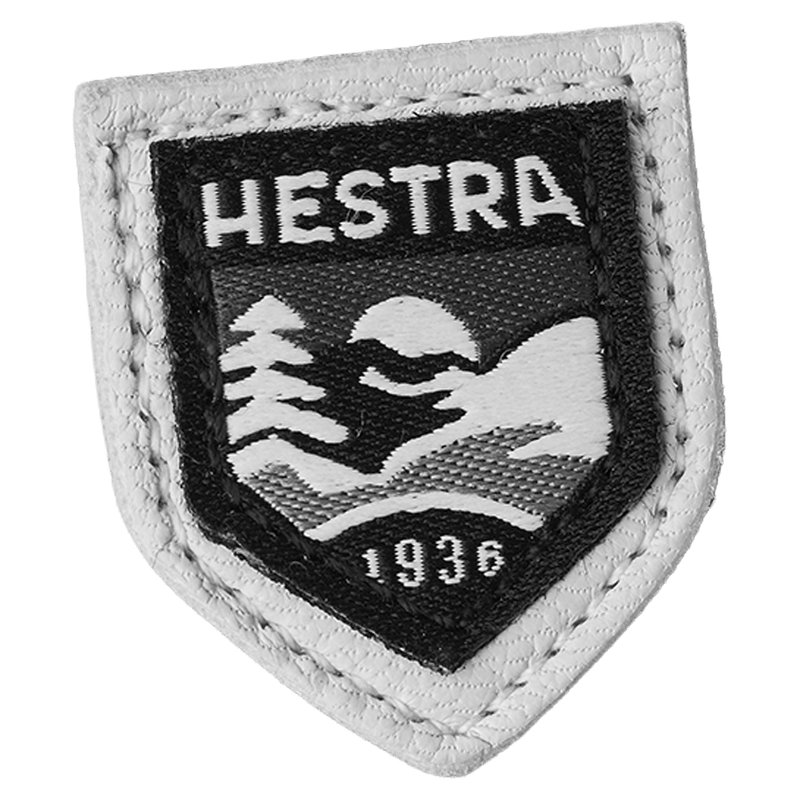 Shield-shaped Hestra logo patch with white leather trim and black embroidered mountain scene and the year 1936.