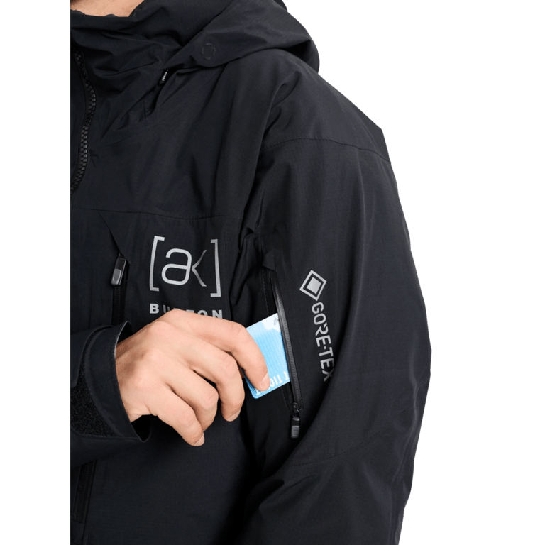 Black Burton AK Lz Gore-Tex jacket with a zippered sleeve pocket and a hand inserting a blue lift pass.
