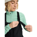 Woman adjusting black velcro shoulder straps on Burton Reserve Bib Snowboard Pants over a mint green base layer.