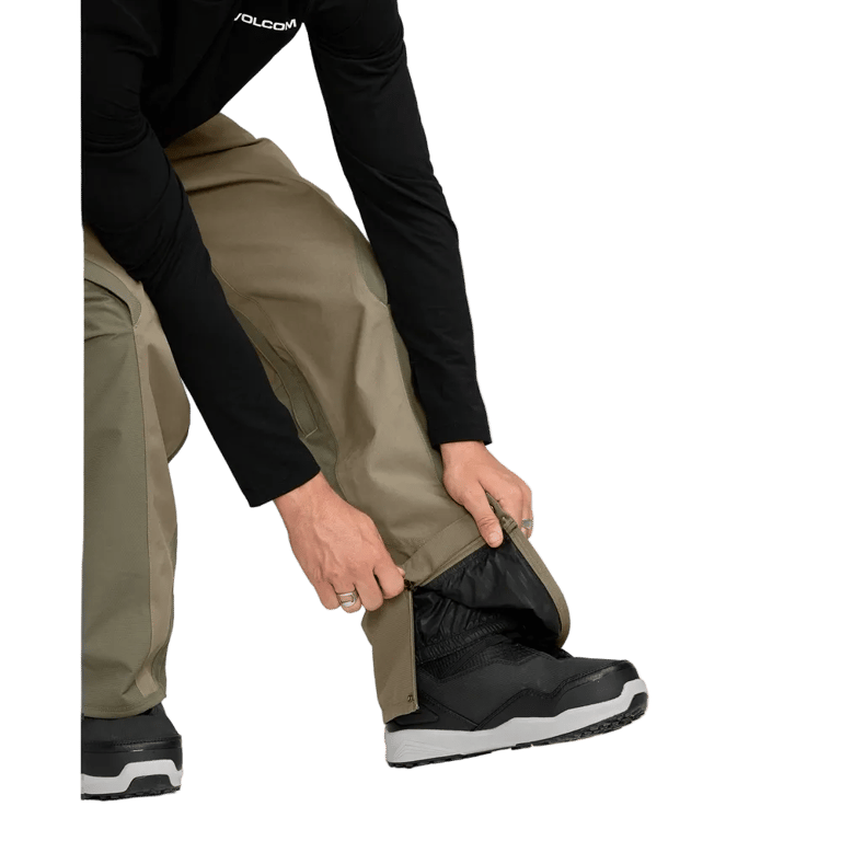 Person adjusting the zippered ankle cuff of olive Volcom Kleveland snowboard pants over a black snowboard boot.