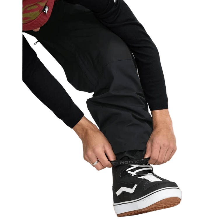 Black Volcom Longo Gore-Tex snowboard pants with elasticated ankle cuffs being adjusted over a black snowboard boot.