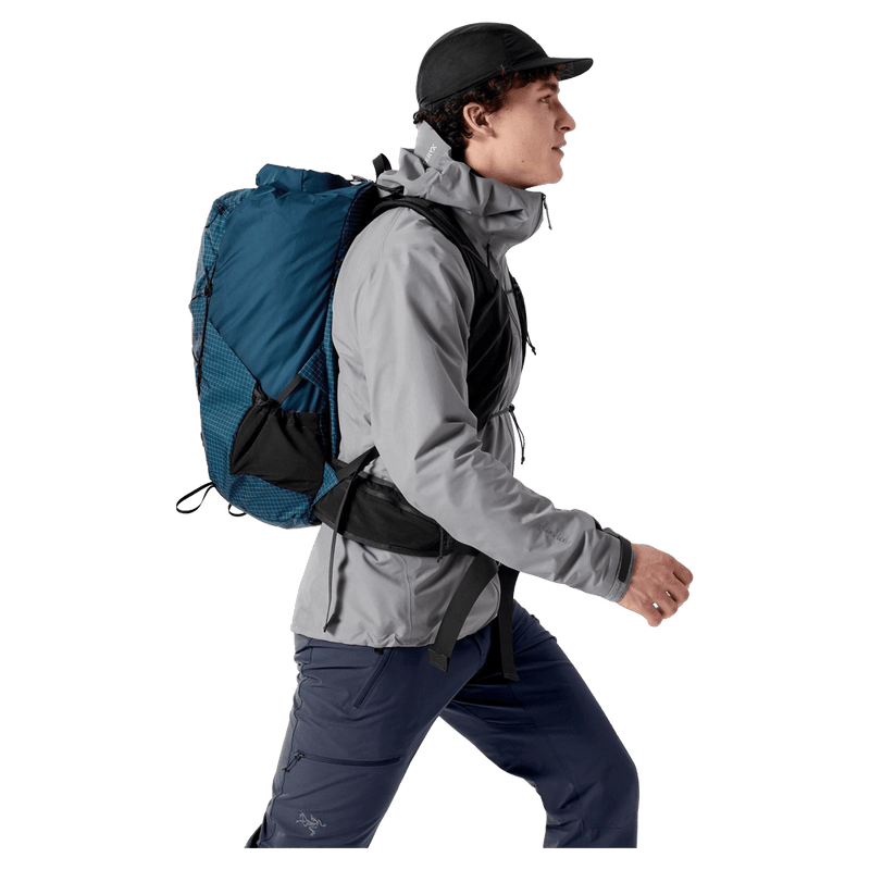 Side profile of a blue Arcteryx Aerios 35 backpack with mesh pockets and waist strap worn by a man walking.