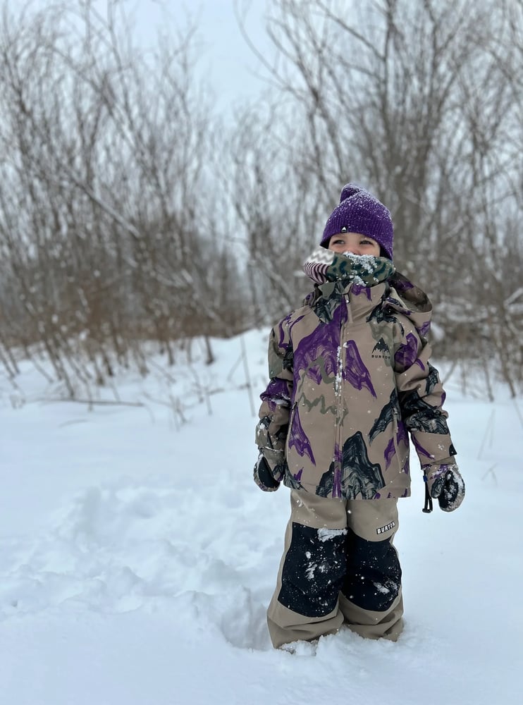 Child wearing tan Burton Maven Bib Snowboard Pants with black knee patches and a patterned jacket in a snowy field.