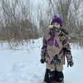 Child wearing tan Burton Maven Bib Snowboard Pants with black knee patches and a patterned jacket in a snowy field.