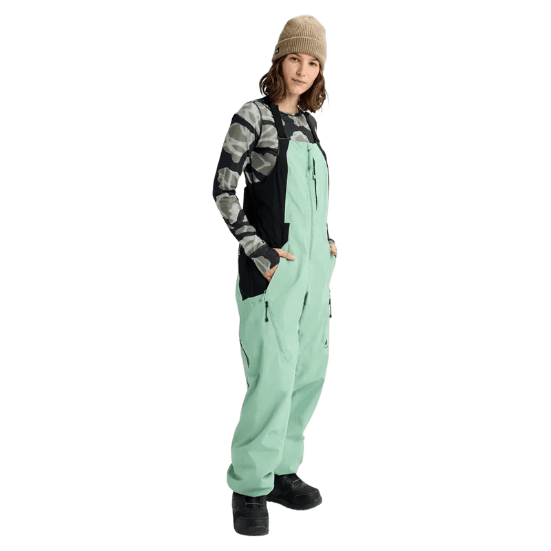 Women's mint green Burton Reserve Bib snowboard pants with black side panels and zippered pockets on a model.