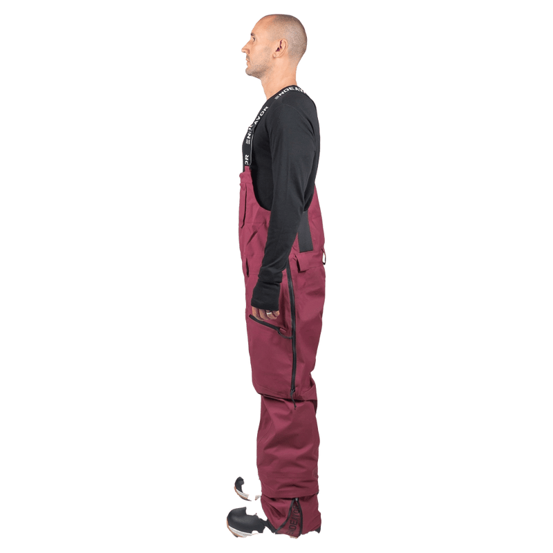 Side profile of a man wearing maroon Endeavor 3L Shelter Bib Snowboard Pants with logo straps and side zippers.