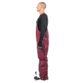 Side profile of a man wearing maroon Endeavor 3L Shelter Bib Snowboard Pants with logo straps and side zippers.