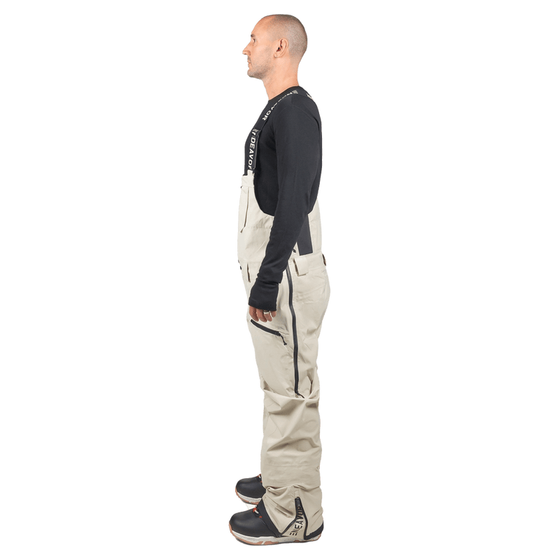 Side profile of beige Endeavor 3L Shelter Bib Snowboard Pants with black side zippers and branded suspender straps.