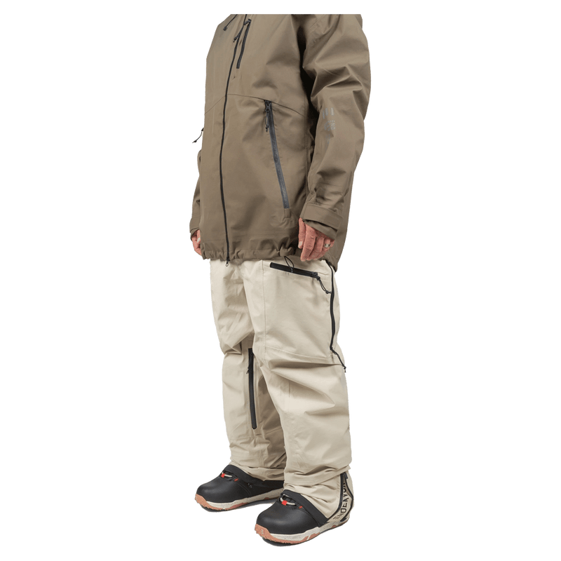 Side profile of beige Endeavor 3L Shelter Bib Snowboard Pants with black zippers, worn with a brown jacket and boots.
