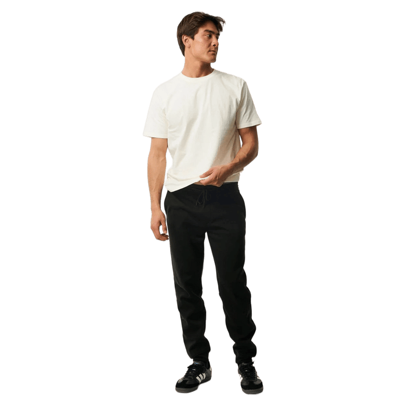 Man wearing black Rip Curl Anti Series Departed Trackpants with a white t-shirt, featuring a tapered leg and cuffed hem.