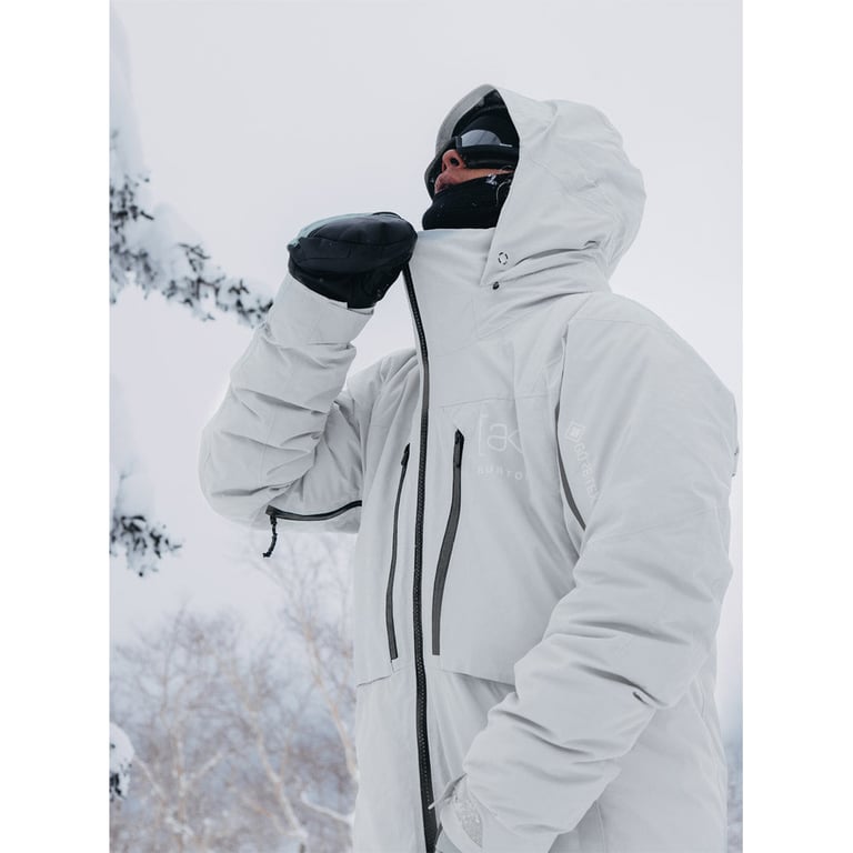 Light grey Burton AK Lz Gore-Tex down snowboard jacket with hood and black zippers worn in a snowy outdoor setting.