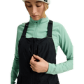 Woman wearing black Burton Reserve Bib Snowboard Pants with a mint green base layer, highlighting the chest zip pocket.