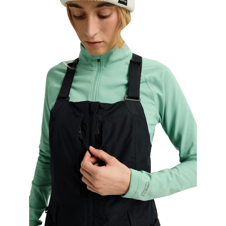 Woman wearing black Burton Reserve Bib Snowboard Pants with a mint green base layer, highlighting the chest zip pocket.