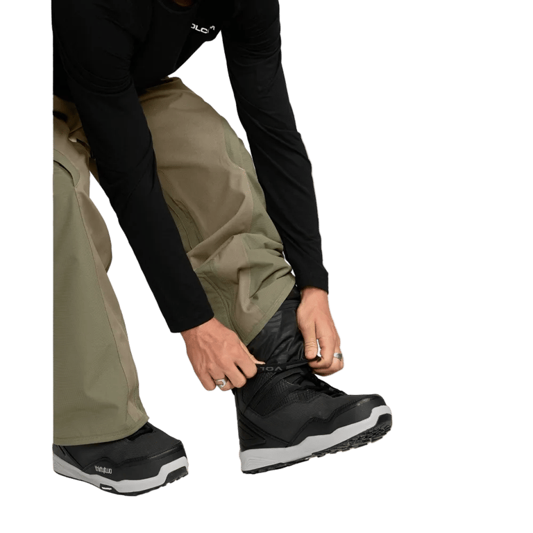 Olive green Volcom Kleveland snowboard pants with adjustable inner gaiter being pulled over black snowboard boots.