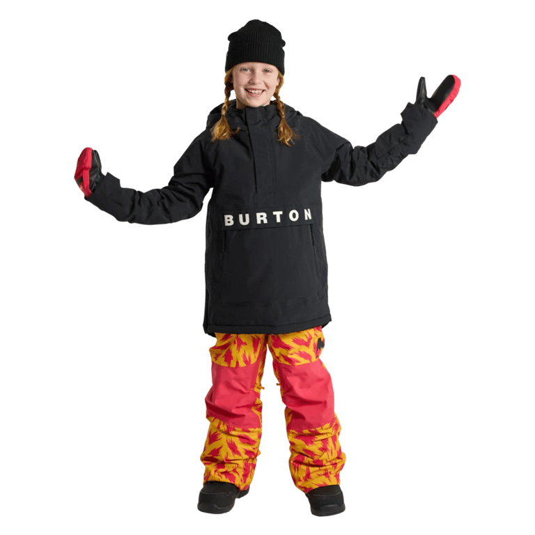 Child wearing a black Burton Frostner Anorak jacket with white logo, black beanie, and patterned snowboard pants.