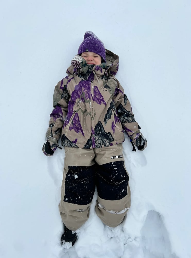 Child wearing tan Burton Maven Bib Snowboard Pants with black knee patches lying in the snow.