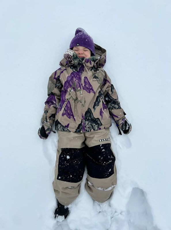 Child wearing tan Burton Maven Bib Snowboard Pants with black knee patches lying in the snow.