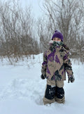 Child wearing tan Burton Maven Bib Snowboard Pants with black knee patches and a patterned jacket in a snowy field.
