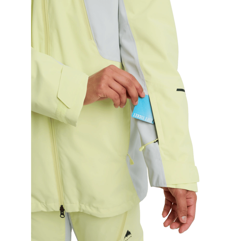 Pale yellow women's Burton Reserve 3-In-1 Snowboard Jacket with a blue lift ticket being placed in a sleeve pocket.