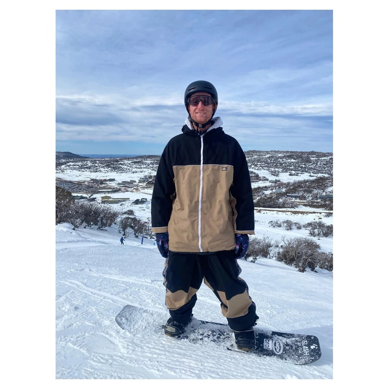 CHECKtheFeed VX Daily Snowboard Jacket in black and tan with white zipper, worn by a snowboarder on a snowy mountain.