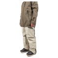 Side profile of beige Endeavor 3L Shelter Bib Snowboard Pants with black zippers, worn with a brown jacket and boots.