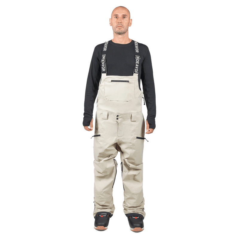 Beige Endeavor 3L Shelter Bib Snowboard Pants with black branded suspenders and zippered pockets on a male model.