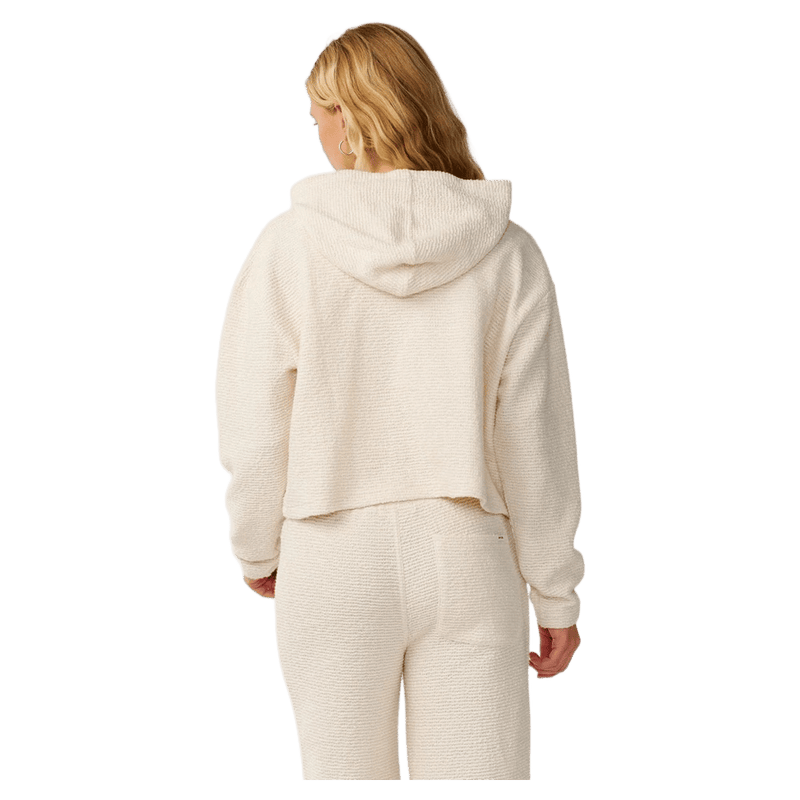 Back view of a woman wearing a cream Rip Curl Cala Nights textured cropped hoodie with a hood.
