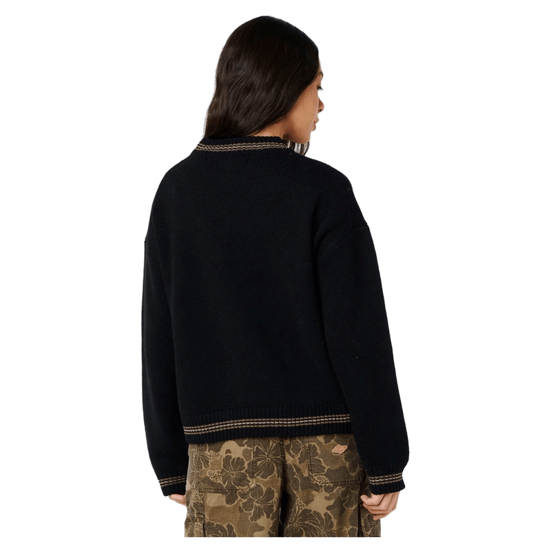 Back view of a woman in a black Rip Curl Half Moon Bay sweater with tan striped trim on the collar, hem, and cuffs.